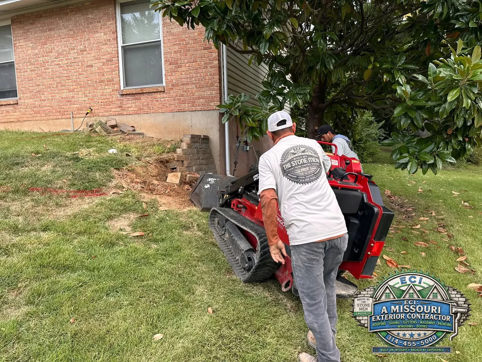 Crew member guiding compact equipment beside a residential retaining-wall site