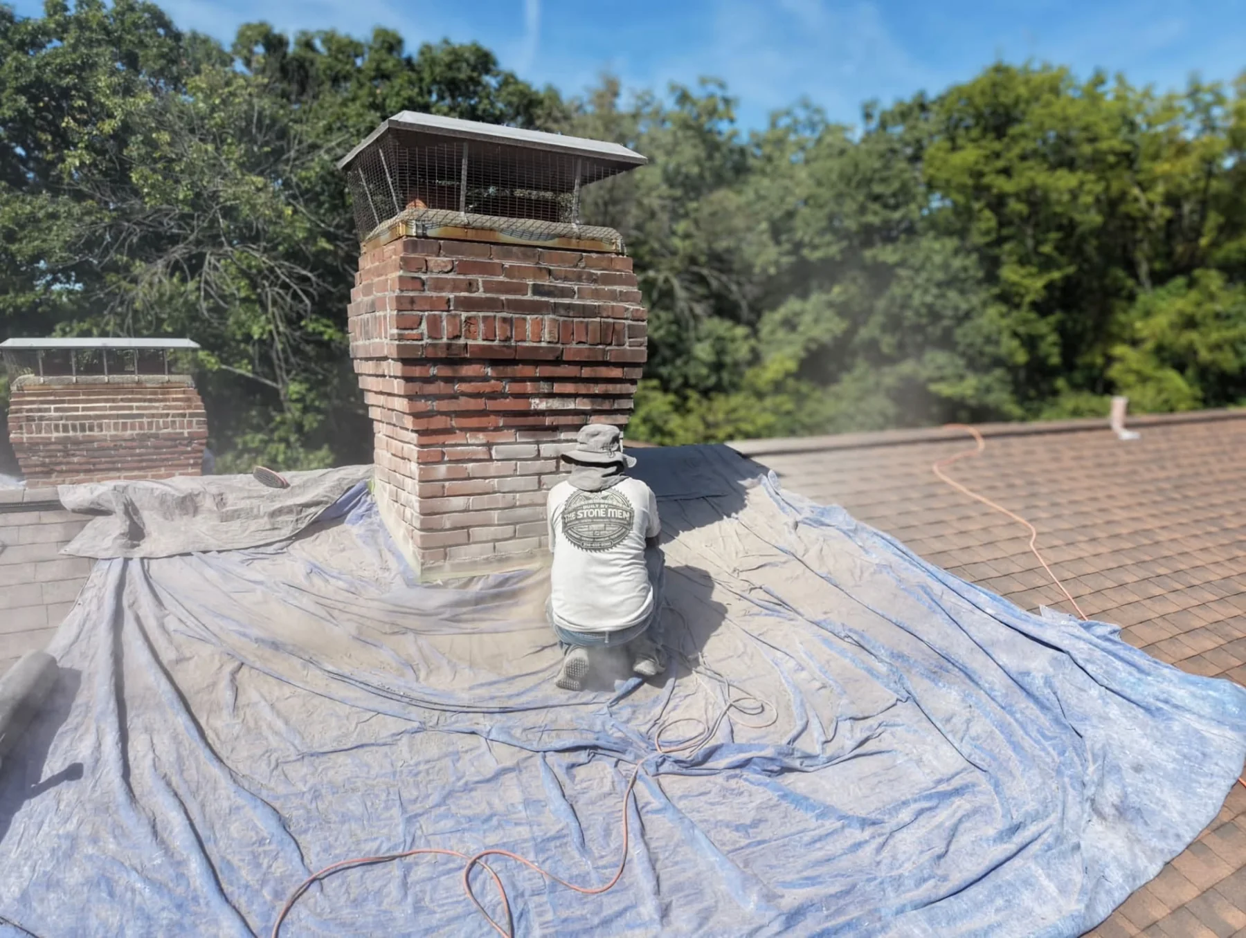 Mason kneeling beside a brick chimney with exposed joints and roof tarps